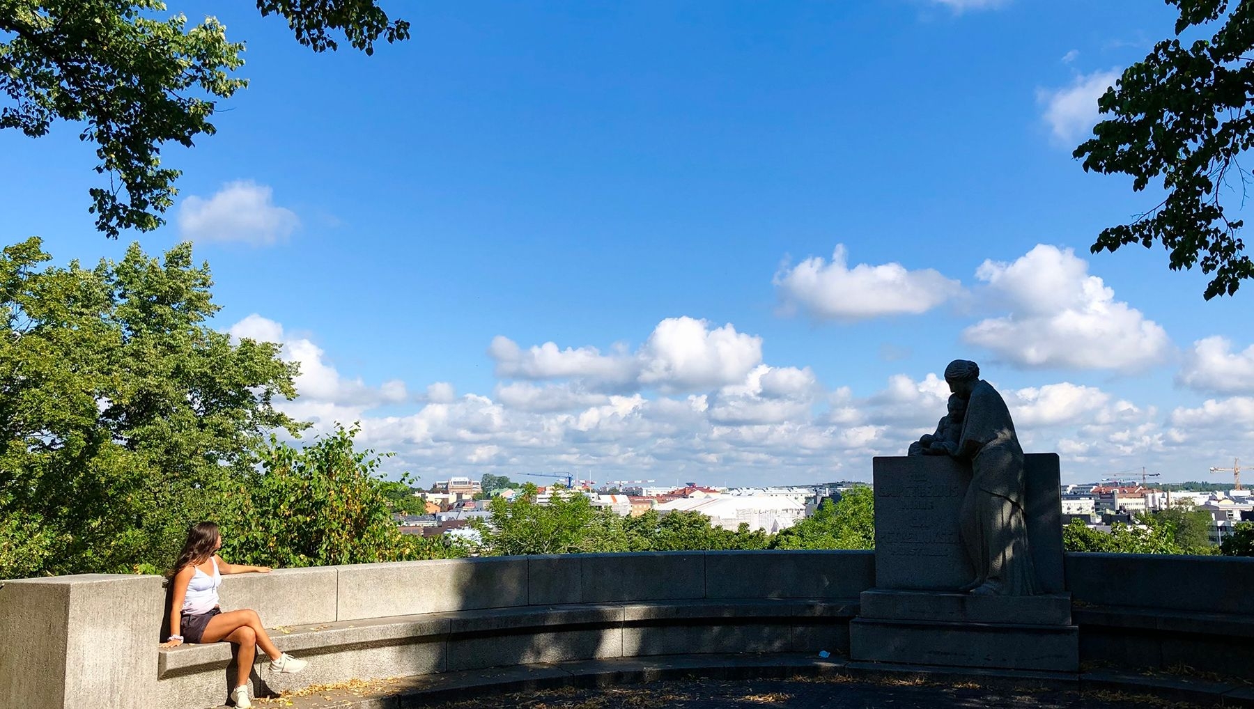 Henkilö istuu G. A. Petreliuksen muistomerkin 11 metriä leveällä betonisella kaarella ja katsoo kohti kaupunkia. Muistomerkki on puiden varjostama. Taivaalla on pilvenhattaroita.