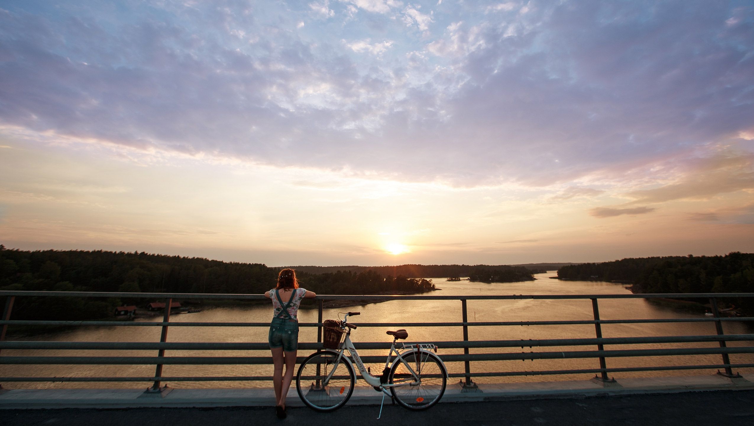 Nainen seisoo sillalla polkupyöränsä vieressä ja nojaa kaiteeseen. Hän katsoo kohti saaristoa ja auringonlaskua, joka on värjännyt meren oranssiksi.