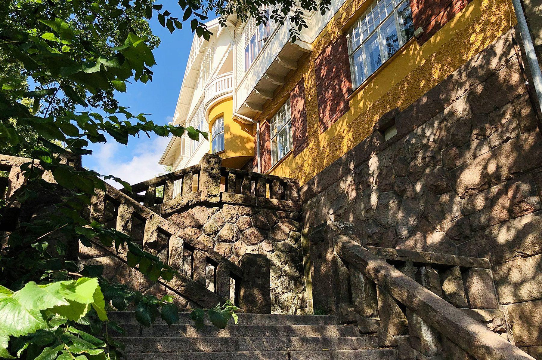 Mustainveljestenkujan mutkittelevat kiviportaat puiden varjossa. Portaiden yläpäässä on keltainen jugend-talo.