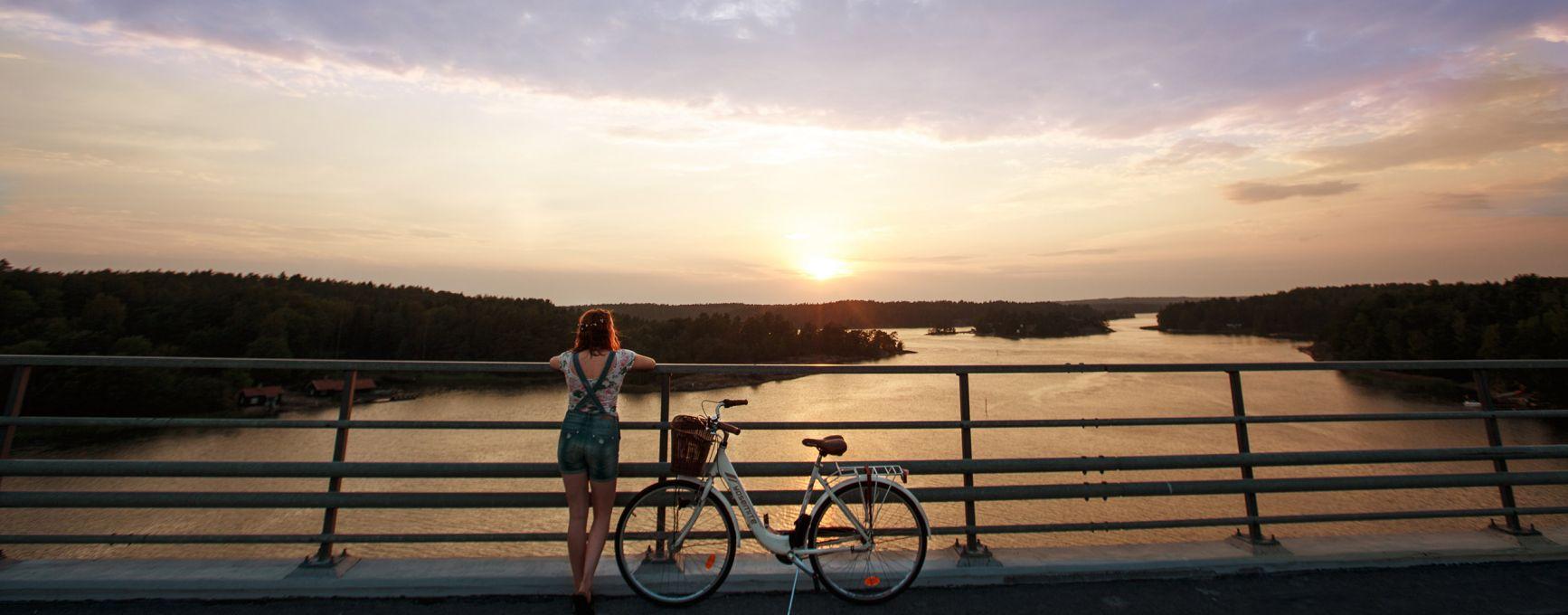 A woman stands, beside her bicycle, on a bridge in the Finnish Archipelago, looking at a sunset.