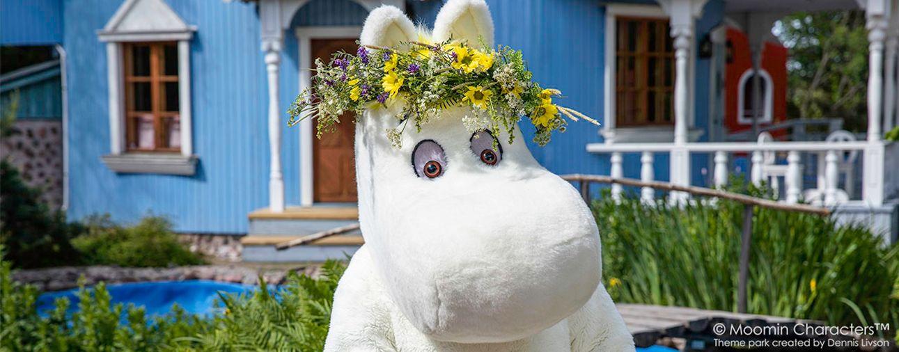 A Moomin character stands in front of the blue Moominhouse at Moominworld.